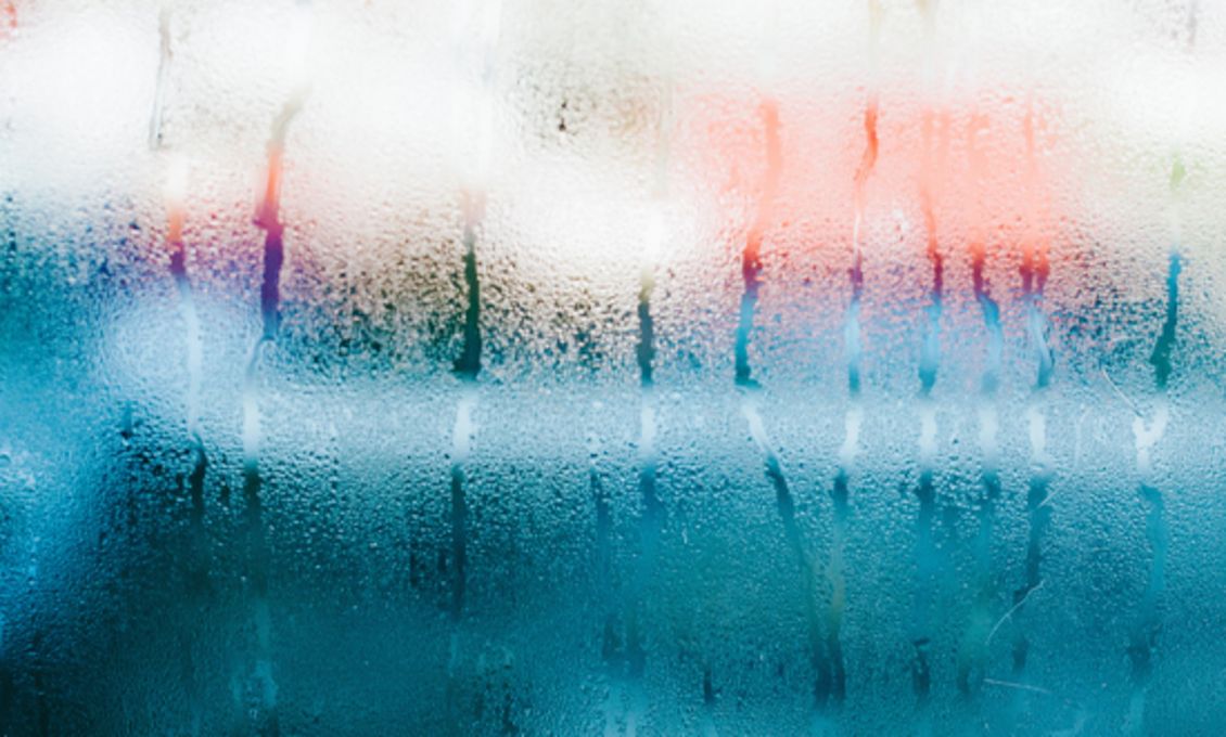 Beschlagene Glasscheibe mit Wassertropfen, die in einem abstrakten Farbmuster aus Blau, Weiß und Rot erscheint.