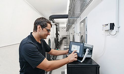 Mitarbeiter der FSM AG bedient ein technisches Gerät in einem Raum mit Belüftungssystemen und Kontrollgeräten an den Wänden.