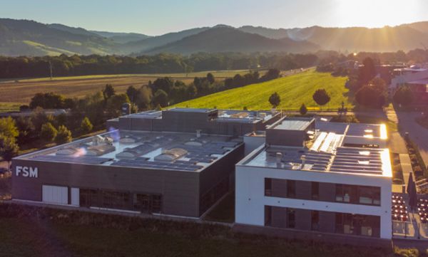 Luftaufnahme des Firmengebäudes der FSM AG bei Sonnenuntergang, umgeben von grüner Landschaft und Hügeln.