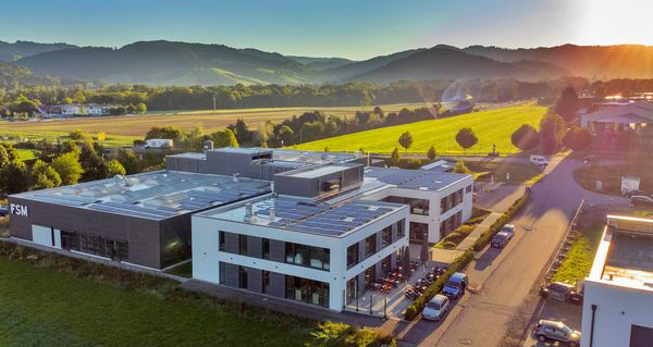 Luftaufnahme des Firmengebäudes der FSM AG bei Sonnenaufgang, umgeben von Feldern, Bäumen und einer ländlichen Landschaft.